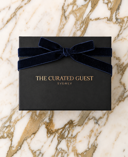 Black gift box with a navy blue ribbon on a marble surface, featuring 'The Curated Guest Sydney'.