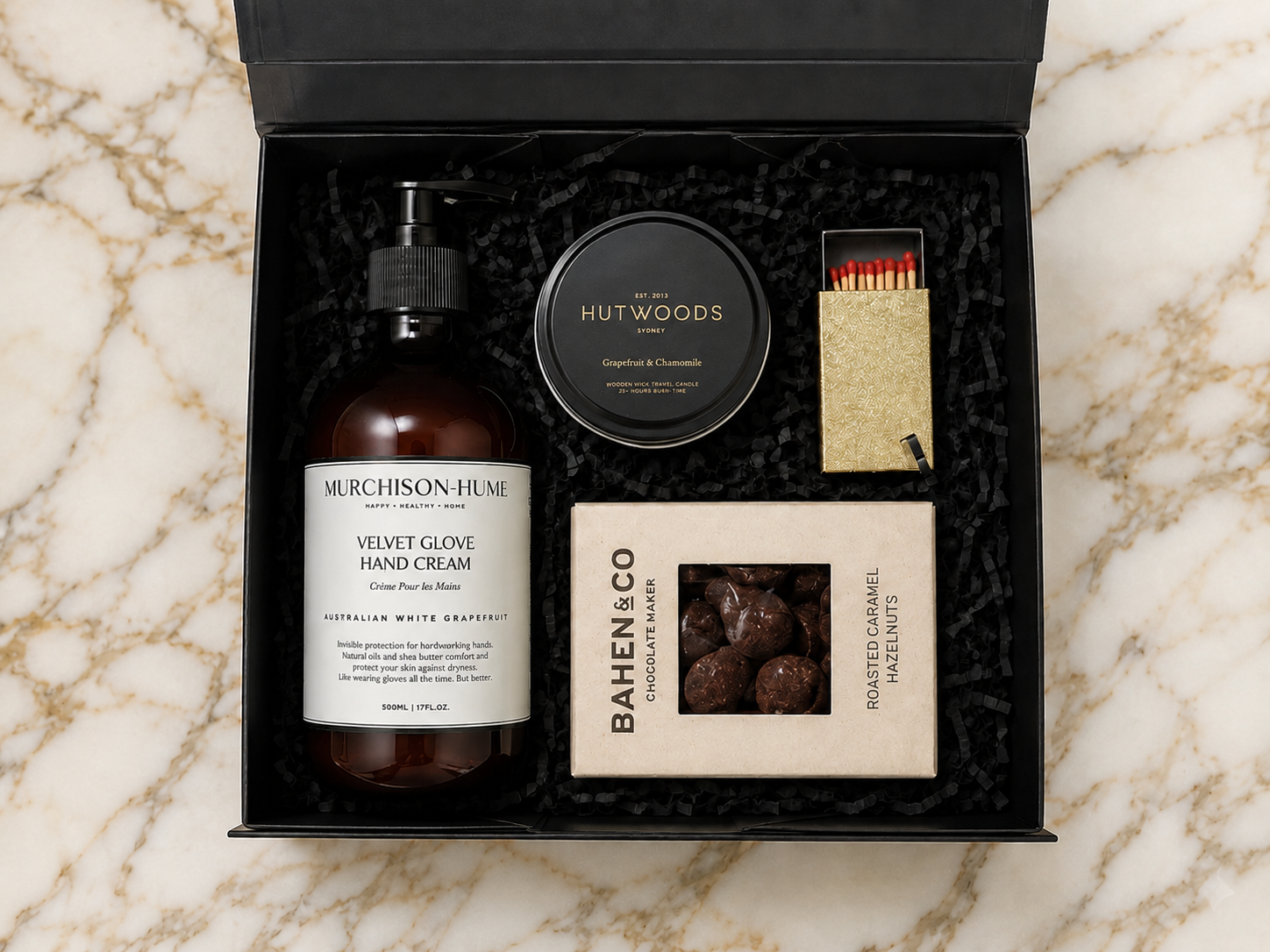 Gift set with Murchison & Hume velvet glove hand cream, Hutwoods travel candle, and Bahen & Co chocolate truffles on a marble surface.